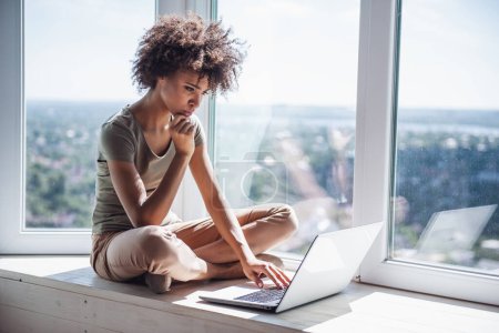 Téléchargez les photos : Belle fille travaille avec un ordinateur portable tout en étant assis sur le rebord de la fenêtre à la maison - en image libre de droit