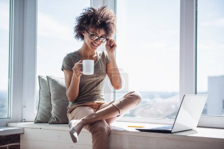 Téléchargez les photos : Belle femme d'affaires utilise un ordinateur portable, boire du café et sourire tout en étant assis sur le rebord de la fenêtre à la maison - en image libre de droit