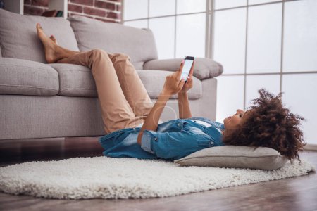 Téléchargez les photos : Belle fille utilise un téléphone intelligent tout en étant couchée sur le sol, ses jambes sur le canapé - en image libre de droit