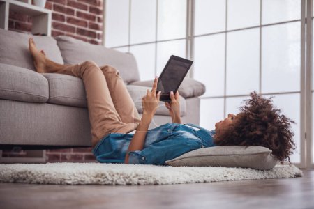 Téléchargez les photos : Belle fille utilise une tablette numérique tout en étant couchée sur le sol, ses jambes sur le canapé - en image libre de droit