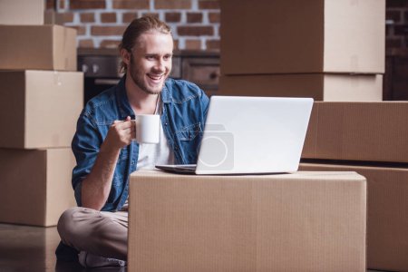 Téléchargez les photos : Beau gars déménage dans un nouvel appartement ; il utilise un ordinateur portable, boire du café et sourire tout en étant assis parmi des boîtes en carton - en image libre de droit