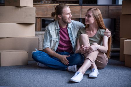 Téléchargez les photos : Beau jeune couple se regarde et sourit tout en étant assis sur le sol parmi les boîtes en carton - en image libre de droit