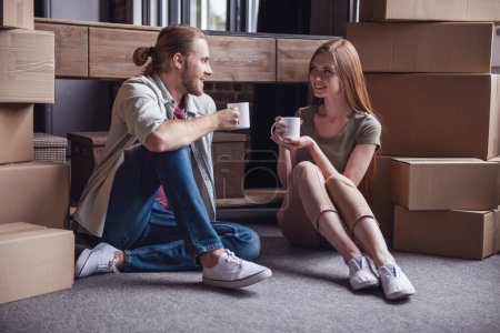 Téléchargez les photos : Beau jeune couple boit du café, parle et sourit tout en étant assis sur le sol parmi les boîtes en carton - en image libre de droit