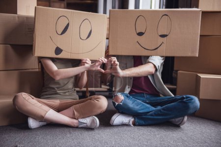 Téléchargez les photos : Beau jeune couple est assis sur le sol de leur nouvel appartement avec des boîtes en carton sur la tête et montrant le cœur - en image libre de droit