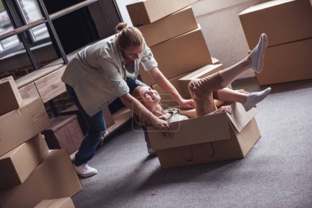 Photo for Beautiful young couple is having fun while moving into new apartment. Girl is sitting in the cardboard boxes - Royalty Free Image