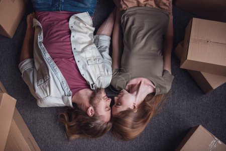 Téléchargez les photos : Vue de dessus de beau jeune couple touchant avec leurs fronts et souriant tout en étant couché sur le sol parmi les boîtes en carton dans leur nouvel appartement - en image libre de droit