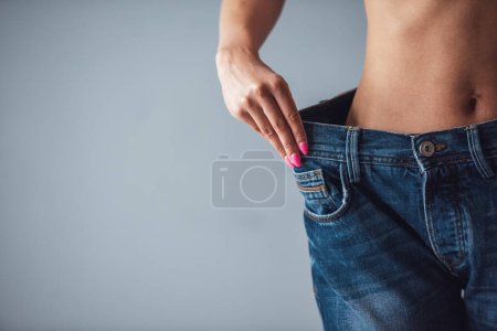 Foto de Imagen recortada de la hermosa chica en pantalones vaqueros de gran tamaño que muestra su pérdida de peso, sobre fondo gris - Imagen libre de derechos