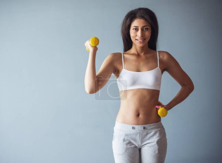 Photo for Beautiful sports girl is holding dumbbells, looking at camera and smiling, on gray background - Royalty Free Image