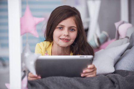 Téléchargez les photos : Petite fille mignonne utilise une tablette numérique, regardant la caméra et souriant tout allongé sur le lit dans sa chambre à la maison - en image libre de droit