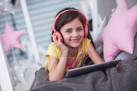 Téléchargez les photos : Petite fille mignonne dans les écouteurs utilise une tablette numérique, regardant la caméra et souriant tout couché sur le lit dans sa chambre à la maison - en image libre de droit