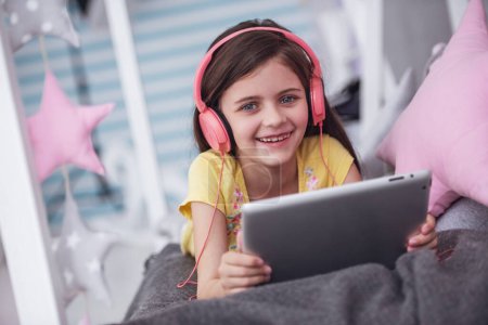 Téléchargez les photos : Petite fille mignonne dans les écouteurs utilise une tablette numérique, regardant la caméra et souriant tout couché sur le lit dans sa chambre à la maison - en image libre de droit