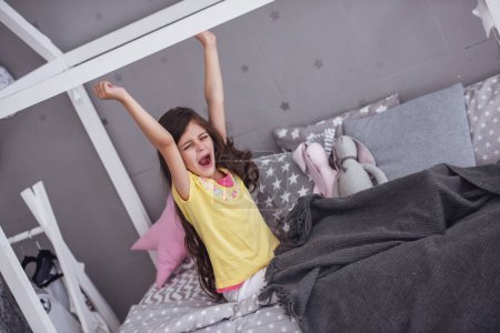 Téléchargez les photos : Petite fille mignonne s'étire et bâille tout en se réveillant dans sa chambre à la maison - en image libre de droit