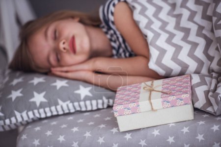 Téléchargez les photos : Jolie petite fille sourit tout en dormant dans sa chambre à la maison, une boîte cadeau est couché près - en image libre de droit