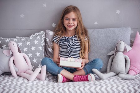 Téléchargez les photos : Jolie petite fille tient une boîte cadeau, regardant la caméra et souriant tout en étant assis sur son lit dans sa chambre à la maison - en image libre de droit