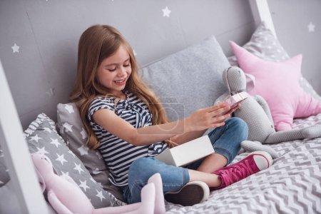 Foto de Linda niña está abriendo una caja de regalo y sonriendo mientras está sentada en su cama en su habitación en casa - Imagen libre de derechos