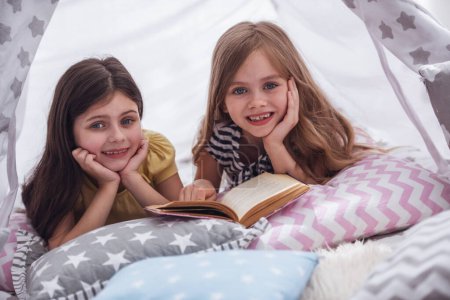 Photo for Two cute little girls are reading a book, looking at camera and smiling while playing together in child's teepee - Royalty Free Image