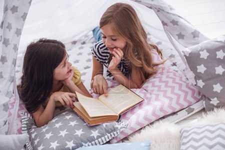 Téléchargez les photos : Deux jolies petites filles lisent un livre et sourient tout en jouant ensemble dans le tipi de l'enfant - en image libre de droit