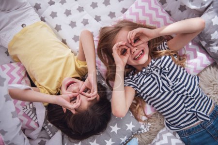 Téléchargez les photos : Vue du dessus de deux jolies petites filles faisant des lunettes avec leurs mains, regardant la caméra et souriant tout en jouant ensemble à la maison - en image libre de droit