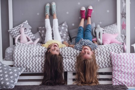 Téléchargez les photos : Deux jolies petites filles regardent la caméra et sourient tout en étant allongées sur le dos sur le lit dans la chambre des enfants - en image libre de droit