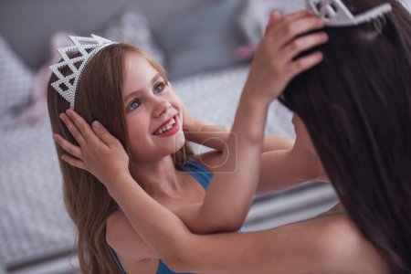 Foto de Dos niñas felices están poniendo coronas en la cabeza del otro y sonriendo mientras juegan en la habitación de los niños en casa - Imagen libre de derechos