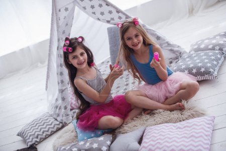 Photo for Two happy little girls in crowns are doing each other hair, looking at camera and smiling while playing in children's room at home - Royalty Free Image