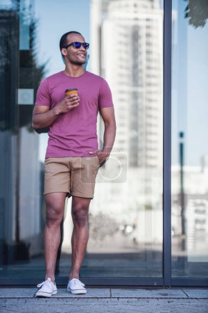 Foto de Retrato de cuerpo entero del hombre guapo sosteniendo una taza de café, mirando hacia otro lado y sonriendo mientras camina por la ciudad - Imagen libre de derechos