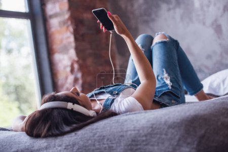 Photo for Beautiful girl in denim overall and headphones is listening to music using a smart phone while lying on bed at home - Royalty Free Image