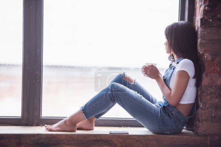 Téléchargez les photos : Belle fille en denim global tient une tasse et regarde par la fenêtre tout en étant assis sur le rebord de la fenêtre à la maison - en image libre de droit