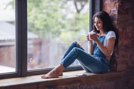 Téléchargez les photos : Belle fille en denim global tient une tasse et souriant tout en étant assis sur le rebord de la fenêtre à la maison - en image libre de droit