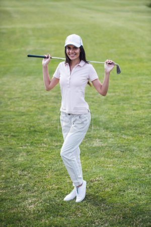 Téléchargez les photos : Portrait complet de belle jeune femme tenant un club de golf, regardant la caméra et souriant tout en se tenant debout sur le terrain de golf - en image libre de droit