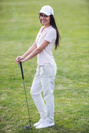 Téléchargez les photos : Portrait complet de belle jeune femme tenant un club de golf, regardant la caméra et souriant tout en se tenant debout sur le terrain de golf - en image libre de droit