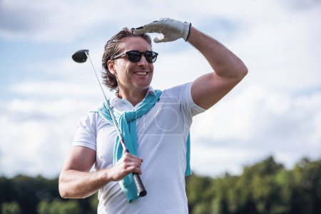 Téléchargez les photos : Beau homme tient un club de golf, regardant loin et souriant tout en se tenant sur le terrain de golf - en image libre de droit