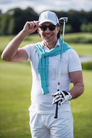 Photo for Handsome man is holding a golf club, looking at camera and smiling while standing on golf course - Royalty Free Image