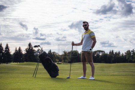 Téléchargez les photos : Portrait complet de beau gars tenant un club de golf et regardant la caméra tout en se tenant debout sur le terrain de golf - en image libre de droit