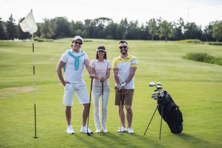 Photo for Two men and a woman are holding golf clubs, looking at camera and smiling while standing on golf course - Royalty Free Image