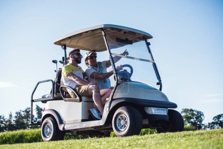 Téléchargez les photos : Beaux hommes parlent et sourient tout en conduisant une voiturette de golf et la recherche d'un trou de golf - en image libre de droit