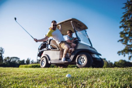 Téléchargez les photos : Les beaux hommes sourient en conduisant une voiturette de golf, un gars élève un club de golf à la balle, balle en évidence - en image libre de droit