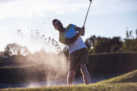 Téléchargez les photos : Beau gars utilise un club de golf tout en jouant au golf, beau soleil brillant, éclaboussé de sable - en image libre de droit