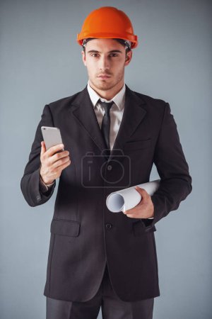 Photo for Handsome young architect in suit and protective helmet is using a smart phone and holding draft, on gray background - Royalty Free Image