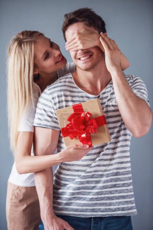 Photo for Beautiful young woman is holding a gift box and covering her boyfriend eyes making a surprise, both are smiling, on gray background - Royalty Free Image