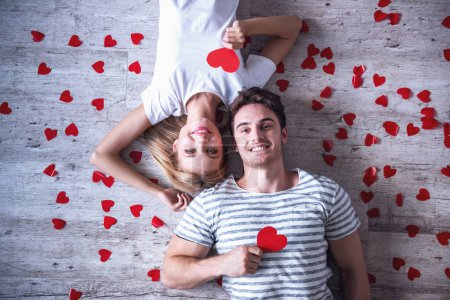 Téléchargez les photos : Vue de dessus de beau jeune couple tenant des coeurs de papier rouge, regardant la caméra et souriant tout en étant couché sur le sol - en image libre de droit