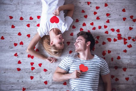 Foto de Vista superior de una hermosa pareja joven sosteniendo corazones de papel rojo, mirándose unos a otros y sonriendo mientras yacen en el suelo - Imagen libre de derechos