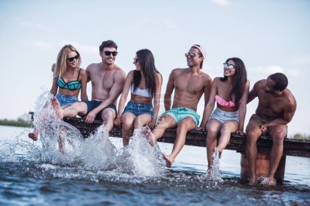 Foto de Felices amigos están salpicando agua y sonriendo mientras están sentados en el muelle en el mar - Imagen libre de derechos
