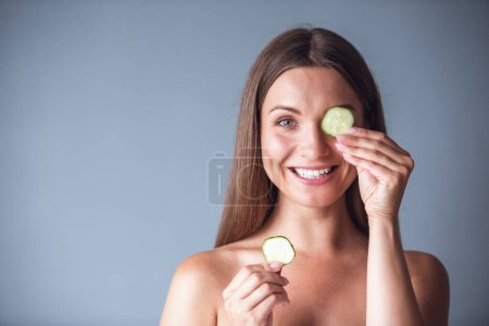 Téléchargez les photos : Belle femme aux épaules nues tient des tranches de concombre, regardant la caméra et souriant, sur fond gris - en image libre de droit