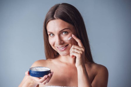 Foto de Hermosa mujer con hombros desnudos está aplicando crema facial, mirando a la cámara y sonriendo, sobre fondo gris - Imagen libre de derechos