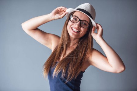 Téléchargez les photos : Belle femme en vêtements décontractés, lunettes et chapeau élégant pose à la caméra et souriant, sur fond gris - en image libre de droit