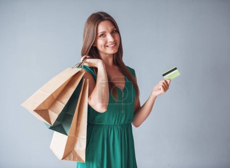 Photo for Beautiful woman in elegant green dress is holding shopping bags and a credit card, looking at camera and smiling, on gray background - Royalty Free Image