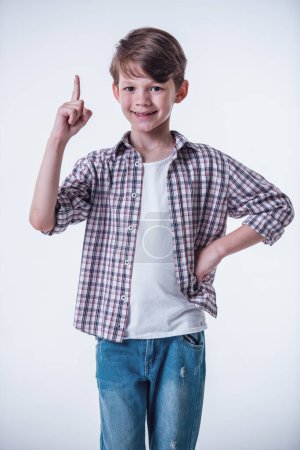 Foto de Niño guapo está mostrando el dedo mientras tiene una idea, mirando a la cámara y sonriendo, sobre un fondo claro - Imagen libre de derechos