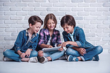 Téléchargez les photos : Les enfants mignons utilisent une tablette numérique et sourient tout en étant assis sur le sol - en image libre de droit