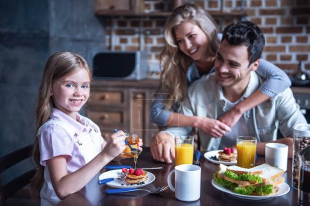 Téléchargez les photos : Joyeux petit déjeuner en famille dans la cuisine. Fille ajoute du miel aux crêpes pendant que les parents la regardent et sourient - en image libre de droit